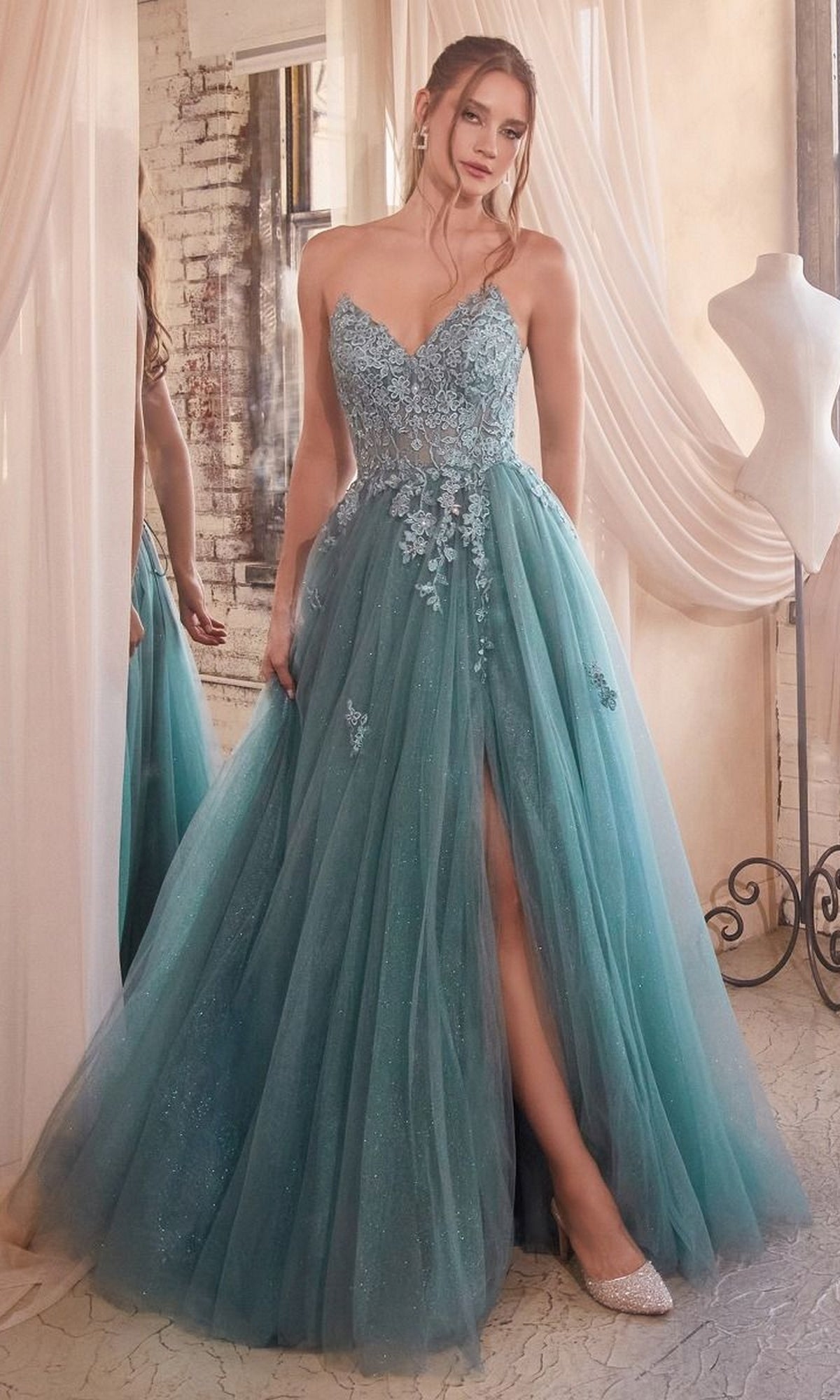 A woman in the Strapless Long Glitter-Tulle Prom Ball Gown C148, featuring a teal strapless design, embroidered lace bodice, floral appliqués, tulle skirt, and thigh-high slit, stands indoors by a mirror and dress form.